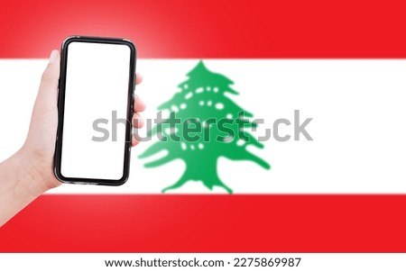 Male hand holding smartphone with blank on screen, on background of blurred flag of Lebanon. Close-up view.