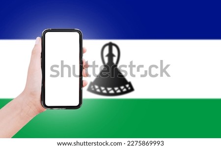 Male hand holding smartphone with blank on screen, on background of blurred flag of Lesotho. Close-up view.