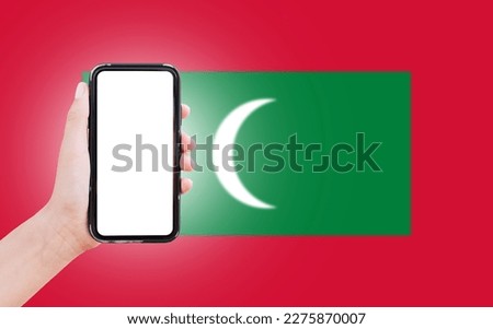 Male hand holding smartphone with blank on screen, on background of blurred flag of Maldives. Close-up view.