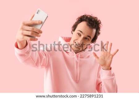 Young man with mobile phone taking selfie on pink background
