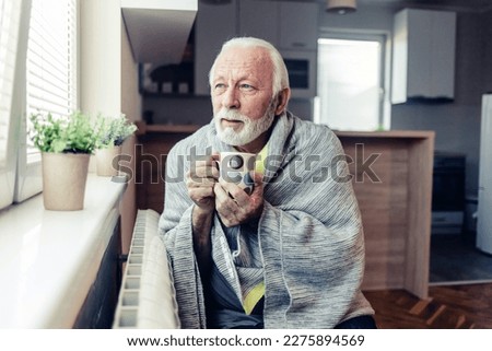Senior man wearing warm clothes is freezing at home and trying to warm up with tea sitting by the radiator. Mature man is feeling cold at home because of an energetic crisis sitting near the radiator.
