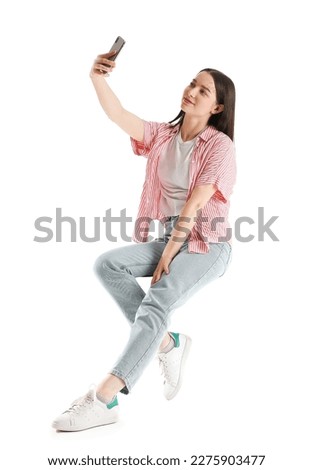 Pretty young woman with smartphone taking selfie while sitting on chair against white background Royalty-Free Stock Photo #2275903477