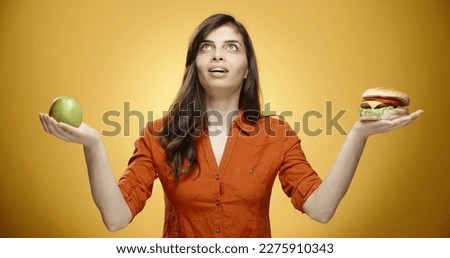 Asian girl in casual clothes suffering from choice between a healthy apple in one hand, and a tasty burger in another hand, isolated on yellow background close up 