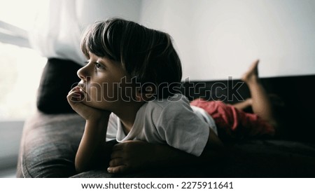 One small boy lying on couch with hand in chin hypnotized by entertainment media at home indoors. Lifestyle shot of kid watching cartoon