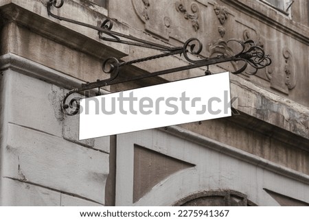 Empty white signboard in vintage metal forged frame hanging on chain on building wall facade. Old fashioned signage with mockup space.