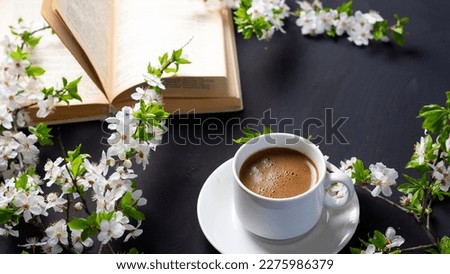 a white cup of espresso coffee, an open book and white cherry blossom branches on a black table.