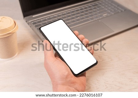 Hand holding mobile phone mockup, smartphone screen at wood desk, laptop. Business app ad.