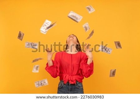 Happy rich young woman under money rain or throwing money around on orange background. Big profit or win lottery concept.