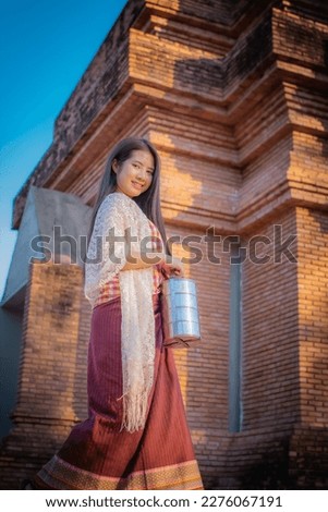 Vertical picture featuring a beautiful thai woman dressed in traditional attire standing in front of an old temple with a smile on her face in the evening light.
