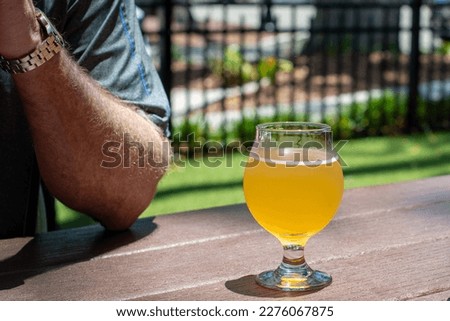 A male reaches for a tall clear glass of sour craft beer in a microbrewery. The cold refreshment has frost on the glass. The beer is on a table and coaster. The top of the glass has white froth. 