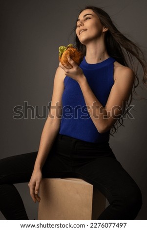 Studio portrait of a beautiful caucasian model in her twenties posing with a cheeseburger. She is wearing black jeans and a blue tanktop. She has brown hair and eyes.