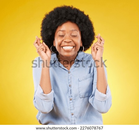 Happy, smile and fingers crossed by black woman in studio for wish, hope and good luck against yellow background. Eyes closed, hand and emoji by excited female waiting, optimistic and hopeful gesture