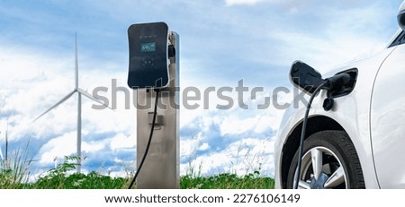 Progressive combination of wind turbine and EV car, future energy infrastructure. Electric vehicle being charged at charging station powered by renewable energy from wind turbine in the countryside. Royalty-Free Stock Photo #2276106149