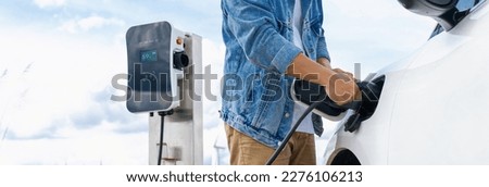 Progressive man with his electric car, EV car recharging energy from charging station on green field with wind turbine as concept of future sustainable energy. Electric vehicle with energy generator. Royalty-Free Stock Photo #2276106213