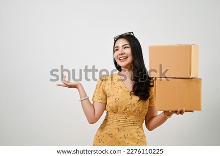 Happy and smiling young Asian woman in casual dress opening her palm and holding cardboard boxes while standing against an isolated white background. shipping service, online shopping concept Royalty-Free Stock Photo #2276110225
