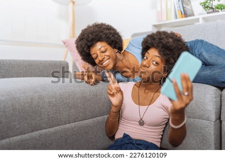 Twin sisters with afro hairstyle and jeans, posing while taking pictures with their light blue cell phone, in the living room of their apartment.