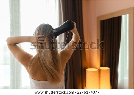 Happy woman drying her hair in bathroom. Hair care. Beautiful smiling woman drying healthy hair using hair dryer. Girl in bath towel is using a dryer and smiling while looking into the mirror. Royalty-Free Stock Photo #2276174377