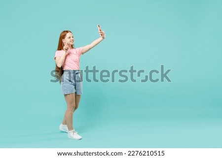 Full length fun little redhead kid girl 12-13 years old in pink striped t-shirt do selfie shot mobile phone post on social network victory gesture isolated on pastel blue background Lifestyle concept