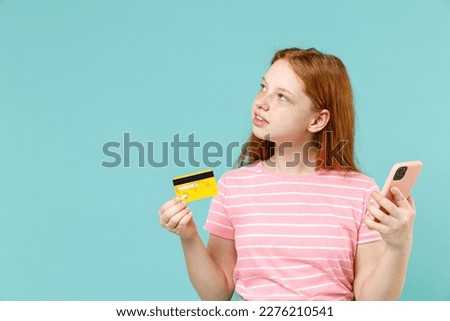 Little wistful redhead kid girl 12-13 years old in pink striped t-shirt hold mobile cell phone credit bank card shopping isolated on pastel blue background studio. Children lifestyle childhood concept