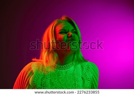 Funny face, grimacing. Young woman emotionally posing over magenta studio background in green neon light. Concept of emotions, facial expression, lifestyle, inspiration, sales, ad
