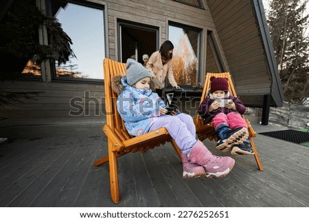 Two little girls sit on chairs at terrace off grid tiny house in the mountains and watching cartoons on mobile phones.