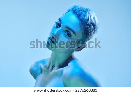 Beautiful young girl with short blonde hair and piercing posing with bare shoulders against blue studio background in neon light. Cyberpunk style. Concept of technology, modern fashion, digital art