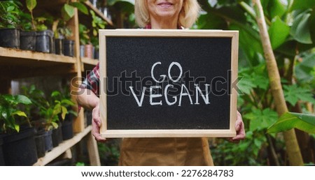 Close up of mature womans hands holding wooden sign go vegan on board, showing to camera in greenhouse behind box with vegetables. Beautiful female biologist posing in hothouse. Hobby farming concept.