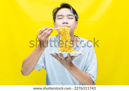 A happy, euphoric Asian young man in a tshirt eating instant fried noodles using bamboo chopstick isolated over a yellow background. stylish asian man with peek a boo hair eats instant noodle