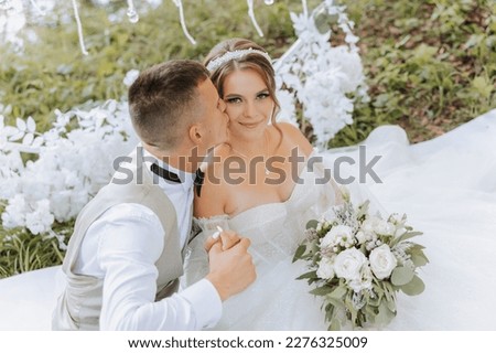The bride and groom on their wedding day, sitting holding hands, kissing her on the cheek outdoors. People are happy and smile. Happy couple in love looking at each other