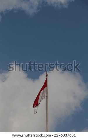 The red and white Indonesian flag flutters in the sky against a background of white clouds and a bright blue sky