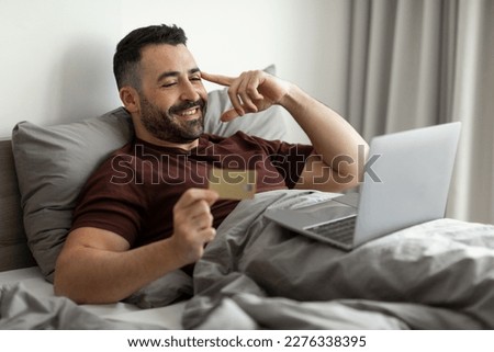 Happy middle aged man using laptop and credit card, sitting in bed at home, shopping online with computer while resting in bedroom, free space