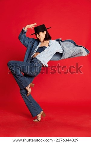 Full-length portrait of beautiful young woman in cowboy style jeans clothes and black hat posing against red studio background. Concept of style, beauty, fashion, youth, emotions. Ad