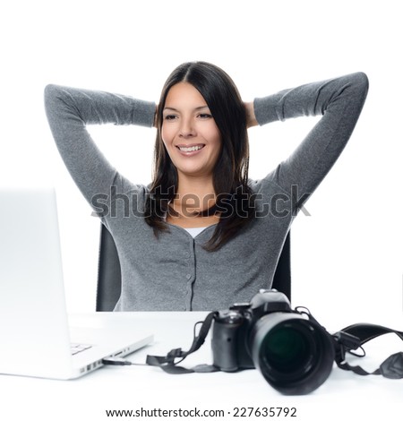 Attractive female photographer sitting back in her chair with her hands clasped behind her head smiling in satisfaction at her captures as she looks at the screen of her laptop with her camera nearby
