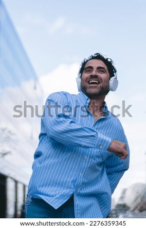 Middle-aged latin man walking happily through the city while wearing headphones. Vertical photo.