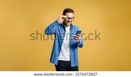 Happy young adult man wearing blue denim shirt adjusting eyeglasses and text messaging over mobile phone isolated on yellow background