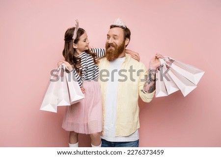 Cheerful girl holding shopping bags and hugging father with crown headband on pink background Royalty-Free Stock Photo #2276373459