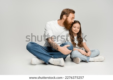 Bearded man in jeans and t-shirt kissing smiling preteen daughter on grey background Royalty-Free Stock Photo #2276375257