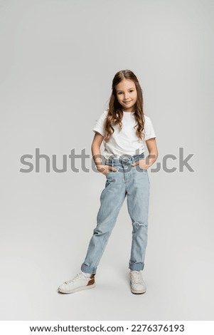 full length of positive child in jeans and t-shirt posing on grey background Royalty-Free Stock Photo #2276376193