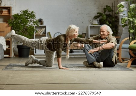 Mature, middle aged woman doing yoga exercises at home with her elderly husband. Couple training together, support and assistance. Concept of family, relationship, retirement, lifestyle, happiness