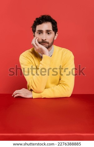 frustrated man in yellow long sleeve jumper sitting at desk on red coral background
