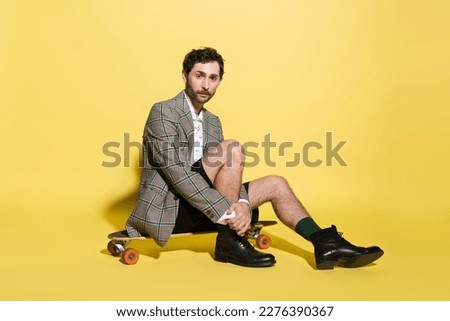 Stylish man in blazer and shorts sitting on skateboard on yellow background