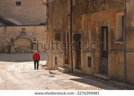 Scanno, L'Aquila, Italy - Picturesque corner of the narrow street with old man with red shirt walking and medieval stone fountain in background in the ancient medieval town. Royalty-Free Stock Photo #2276390481