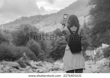 a young tourist woman with beautiful wavy hair, with a backpack on her shoulders, travels through the mountains near the river and photographs nature on her phone, black and white photo.
