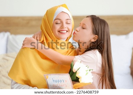 Little girl kissing her Muslim mother with greeting card and tulips in bedroom