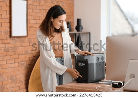 Young woman with portable power station at table in office Royalty-Free Stock Photo #2276413423