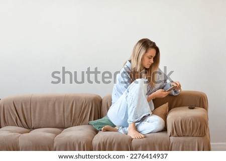 Young blonde woman sitting at home on a couch, lost in the endless scroll of her smartphone screen. Addictive nature of social media and the digital world has taken hold, making it hard to disconnect. Royalty-Free Stock Photo #2276414937