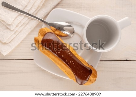 One chocolate eclair with white ceramic saucer, cup and metal spoon on wooden table, close-up, top view. Royalty-Free Stock Photo #2276438909