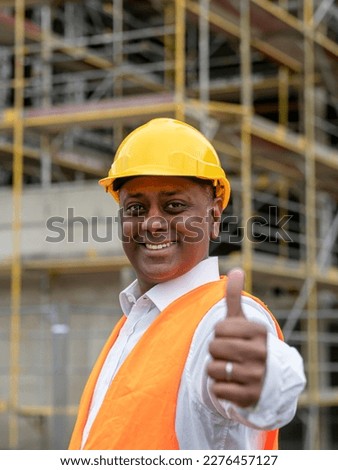 Smiling Indian construction worker wearing safety helmet and vest posing on construction site showing thumbs up gesture