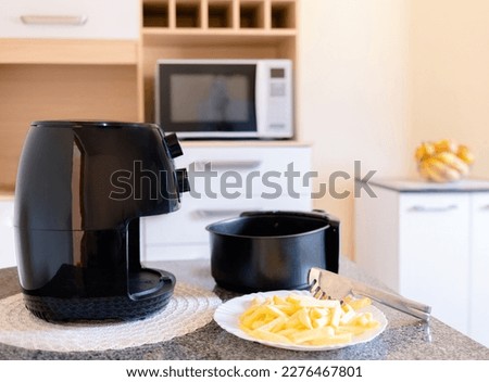 Air fryer on a kitchen table next to a plate of sliced potatoes. Preparing frozen potatoes to fry them in the Air Fryer. Royalty-Free Stock Photo #2276467801