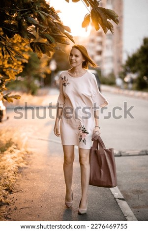 An adult pretty woman of middle age in a dress with a big bag in her hands in the rays of the sunset on a city street. Autumn mood.